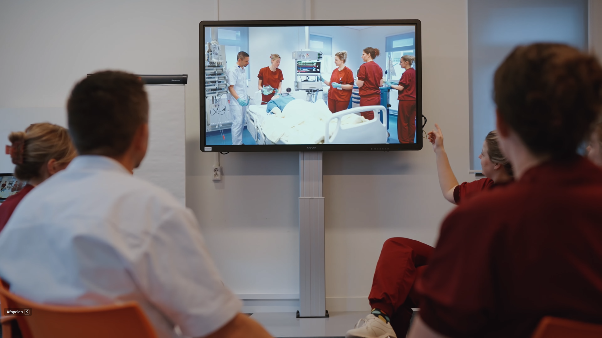 Studenten kijken in een leslokaal naar videobeelden van een simulatietraining op een groot scherm.